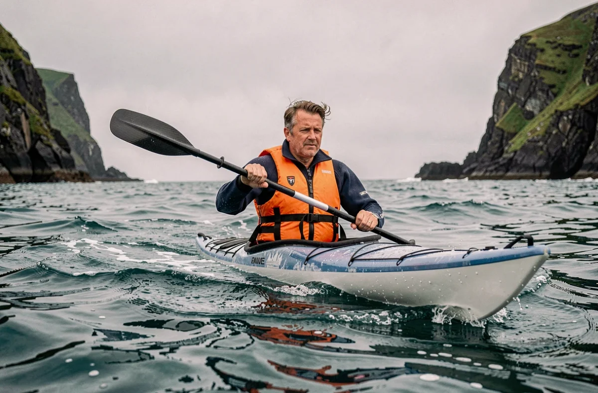 Kayakiste en mer pagayant face aux vagues avec des falaises côtières verdoyantes en arrière-plan