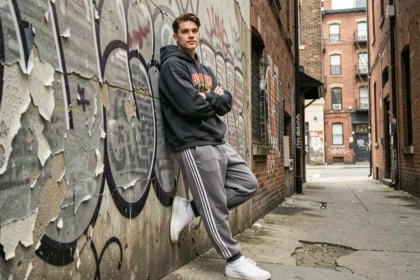 Jeune homme en streetwear portant un hoodie oversize et des sneakers blanches appuyé contre un mur urbain en béton